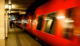 Ein roter Zug rast mit verschwommener Bewegung durch eine schwach beleuchtete U-Bahn-Station. Ein Bahnsteig mit gelben taktilen Markierungen ist sichtbar, in der Ferne steht eine Person. Die Decke ist mit linearen Lichtern beleuchtet.