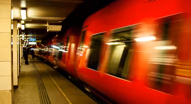 Ein roter Zug rast mit verschwommener Bewegung durch eine schwach beleuchtete U-Bahn-Station. Ein Bahnsteig mit gelben taktilen Markierungen ist sichtbar, in der Ferne steht eine Person. Die Decke ist mit linearen Lichtern beleuchtet.