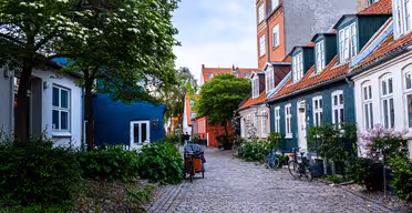 Eine malerische Kopfsteinpflasterstraße mit charmanten Häusern und farbenfrohen Fassaden. Fahrräder parken neben den Gebäuden, und üppige Bäume und Pflanzen schaffen eine ruhige Atmosphäre. Ein blaues Haus und traditionelle Häuser mit roten Dächern verleihen Charakter.