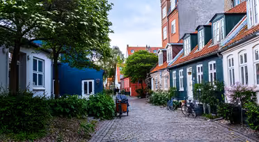 Eine malerische Kopfsteinpflasterstraße mit charmanten Häusern und farbenfrohen Fassaden. Fahrräder parken neben den Gebäuden, und üppige Bäume und Pflanzen schaffen eine ruhige Atmosphäre. Ein blaues Haus und traditionelle Häuser mit roten Dächern verleihen Charakter.