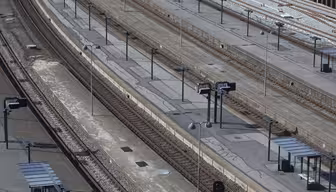 Luftaufnahme eines leeren Bahnhofs mit mehreren parallelen Gleisen und Bahnsteigen. Die Bahnsteige sind mit Schildern und überdachten Wartebereichen gesäumt. Der Bereich ist menschenleer und zugfrei, was die Infrastruktur des Bahnhofs hervorhebt.