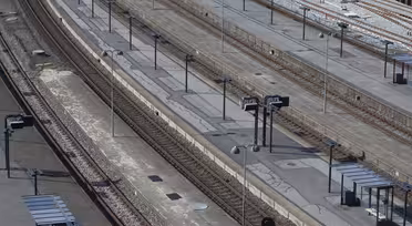 Luftaufnahme eines leeren Bahnhofs mit mehreren parallelen Gleisen und Bahnsteigen. Die Bahnsteige sind mit Schildern und überdachten Wartebereichen gesäumt. Der Bereich ist menschenleer und zugfrei, was die Infrastruktur des Bahnhofs hervorhebt.