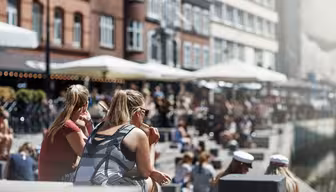 An einem sonnigen Tag sitzen Menschen an einem Kanal mit Backsteingebäuden und Straßencafés am Wasser. Die Szene ist lebhaft, die Leute genießen das Wetter, manche tragen Sonnenbrillen, andere weiße Mützen.