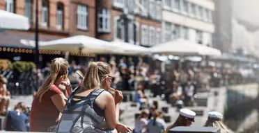 An einem sonnigen Tag sitzen Menschen an einem Kanal mit Backsteingebäuden und Straßencafés am Wasser. Die Szene ist lebhaft, die Leute genießen das Wetter, manche tragen Sonnenbrillen, andere weiße Mützen.