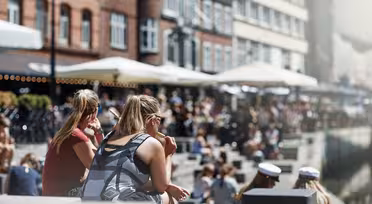 An einem sonnigen Tag sitzen Menschen an einem Kanal mit Backsteingebäuden und Straßencafés am Wasser. Die Szene ist lebhaft, die Leute genießen das Wetter, manche tragen Sonnenbrillen, andere weiße Mützen.