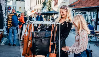 Zwei Frauen stöbern auf einem Markt im Freien nach Ledertaschen. Die Straße ist von Gebäuden gesäumt, im Hintergrund sind weitere Käufer und Verkäufer zu sehen. Die Szene ist lebhaft und farbenfroh.
