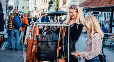 Zwei Frauen stöbern auf einem Markt im Freien nach Ledertaschen. Die Straße ist von Gebäuden gesäumt, im Hintergrund sind weitere Käufer und Verkäufer zu sehen. Die Szene ist lebhaft und farbenfroh.