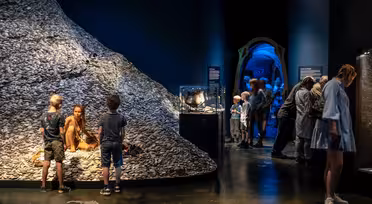 Besucher erkunden eine schwach beleuchtete Ausstellung in einem Museum. Eine Frau sitzt neben einem mit Muscheln bedeckten Hügel und unterhält sich mit Kindern. In der Nähe betrachten Menschen Schautafeln und Informationstafeln entlang eines dunklen, blau beleuchteten Korridors.