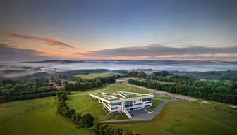 Ein modernes, eckiges Gebäude ist von weitläufigen grünen Feldern und Bäumen umgeben. Eine Nebelschicht schwebt über der Landschaft, ein farbenfroher Sonnenaufgang erhellt den Horizont und vereinzelte Wolken. Eine Auffahrt führt zum Gebäude, das vor sanften Hügeln liegt.
