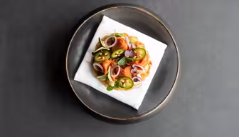 Ein Gourmetgericht mit einem runden Gebäckstück, belegt mit grünen Jalapeño-Scheiben, roten Zwiebeln und garniert mit Kräutern. Die Backwaren liegen auf einer weißen Serviette, die in der Mitte eines dunklen runden Tellers auf dunklem Hintergrund platziert ist.