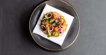 Ein Gourmetgericht mit einem runden Gebäckstück, belegt mit grünen Jalapeño-Scheiben, roten Zwiebeln und garniert mit Kräutern. Die Backwaren liegen auf einer weißen Serviette, die in der Mitte eines dunklen runden Tellers auf dunklem Hintergrund platziert ist.