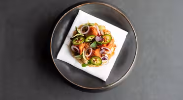 Ein Gourmetgericht mit einem runden Gebäckstück, belegt mit grünen Jalapeño-Scheiben, roten Zwiebeln und garniert mit Kräutern. Die Backwaren liegen auf einer weißen Serviette, die in der Mitte eines dunklen runden Tellers auf dunklem Hintergrund platziert ist.