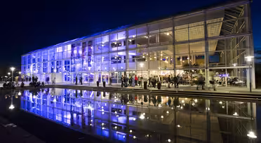 Ein modernes Glasgebäude mit leuchtend blauer und violetter Beleuchtung spiegelt sich nachts in einem großen Wasserbecken. Draußen haben sich Menschen versammelt, was für eine lebhafte Atmosphäre sorgt.