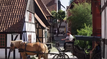 Eine Pferdekutsche fährt durch eine Kopfsteinpflasterstraße in einem malerischen Dorf mit Fachwerkhäusern. Im Hintergrund ist eine Windmühle zu sehen. Ein Kutscher lenkt das Pferd, und ein Passagier sitzt in der Kutsche.