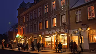 Eine Gruppe von Menschen geht in der Abenddämmerung eine schwach beleuchtete Straße entlang. Die Gebäude sind mit warmen Lichtern und festlichen Girlanden geschmückt. Große Fenster präsentieren verschiedene Waren, und der tiefblaue Himmel schafft eine gemütliche, einladende Atmosphäre.