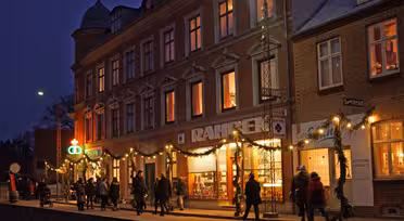 Eine Gruppe von Menschen geht in der Abenddämmerung eine schwach beleuchtete Straße entlang. Die Gebäude sind mit warmen Lichtern und festlichen Girlanden geschmückt. Große Fenster präsentieren verschiedene Waren, und der tiefblaue Himmel schafft eine gemütliche, einladende Atmosphäre.