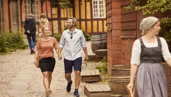 Ein Paar geht Hand in Hand auf einem Kopfsteinpflasterweg, lächelt und genießt einen sonnigen Tag. Der Bereich ist gesäumt von rustikalen Holzhäusern. Im Vordergrund geht eine Frau in historischer Kleidung. Im Hintergrund sind weitere Personen zu sehen.