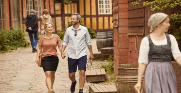 Ein Paar geht Hand in Hand auf einem Kopfsteinpflasterweg, lächelt und genießt einen sonnigen Tag. Der Bereich ist gesäumt von rustikalen Holzhäusern. Im Vordergrund geht eine Frau in historischer Kleidung. Im Hintergrund sind weitere Personen zu sehen.