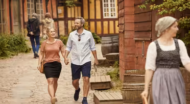 Ein Paar geht Hand in Hand auf einem Kopfsteinpflasterweg, lächelt und genießt einen sonnigen Tag. Der Bereich ist gesäumt von rustikalen Holzhäusern. Im Vordergrund geht eine Frau in historischer Kleidung. Im Hintergrund sind weitere Personen zu sehen.