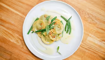 Ein weißer Teller mit ordentlich angerichteten Ravioli, garniert mit Pinienkernen und frischem Basilikum. Grüne Bohnen und eine leichte Sahnesauce begleiten das Gericht, das auf einem Holztisch präsentiert wird.