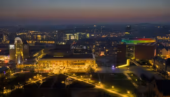 Luftaufnahme von Aarhus bei Nacht. Die Gebäude sind beleuchtet, darunter ein Gebäude mit einem regenbogenfarbenen, runden Dach. Straßen und Wege sind vor einem dunklen Himmel erleuchtet, in der Ferne sind die Lichter der Stadt zu sehen.