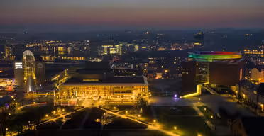 Luftaufnahme von Aarhus bei Nacht. Die Gebäude sind beleuchtet, darunter ein Gebäude mit einem regenbogenfarbenen, runden Dach. Straßen und Wege sind vor einem dunklen Himmel erleuchtet, in der Ferne sind die Lichter der Stadt zu sehen.
