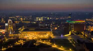 Luftaufnahme von Aarhus bei Nacht. Die Gebäude sind beleuchtet, darunter ein Gebäude mit einem regenbogenfarbenen, runden Dach. Straßen und Wege sind vor einem dunklen Himmel erleuchtet, in der Ferne sind die Lichter der Stadt zu sehen.