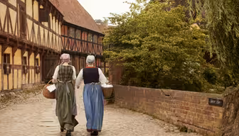 Zwei Personen in historischer Kleidung gehen einen gepflasterten Weg entlang und tragen jeweils einen Weidenkorb. Sie werden von Fachwerkhäusern und einer Backsteinmauer flankiert, auf der rechten Seite befindet sich Grünfläche.