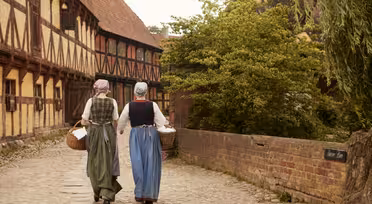 Zwei Personen in historischer Kleidung gehen einen gepflasterten Weg entlang und tragen jeweils einen Weidenkorb. Sie werden von Fachwerkhäusern und einer Backsteinmauer flankiert, auf der rechten Seite befindet sich Grünfläche.