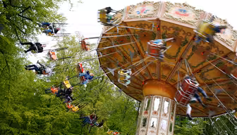 Ein sich drehendes Karussell in einem Freizeitpark, auf dem mehrere Personen sitzen und herumwirbeln. Das Karussell hat ein dekoratives Dach und steht vor einer Kulisse aus üppigen grünen Bäumen. Die Bewegung erzeugt einen Unschärfeeffekt auf den Fahrgästen.