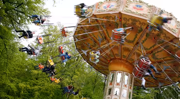 Ein sich drehendes Karussell in einem Freizeitpark, auf dem mehrere Personen sitzen und herumwirbeln. Das Karussell hat ein dekoratives Dach und steht vor einer Kulisse aus üppigen grünen Bäumen. Die Bewegung erzeugt einen Unschärfeeffekt auf den Fahrgästen.