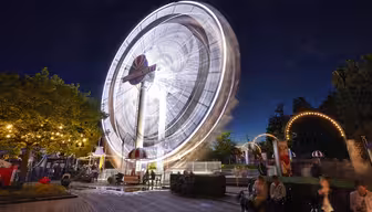 Ein hell erleuchtetes Riesenrad dreht sich nachts in einem Vergnügungspark. Die Bewegung des Rades erzeugt einen verschwommenen Effekt. Menschen sitzen und stehen in der Nähe unter beleuchteten Bäumen und Bögen und genießen die lebhafte Atmosphäre vor einem tiefblauen Himmel.