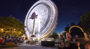 Ein hell erleuchtetes Riesenrad dreht sich nachts in einem Vergnügungspark. Die Bewegung des Rades erzeugt einen verschwommenen Effekt. Menschen sitzen und stehen in der Nähe unter beleuchteten Bäumen und Bögen und genießen die lebhafte Atmosphäre vor einem tiefblauen Himmel.