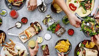 Ein Tisch voller verschiedener Tex-Mex-Gerichte, darunter Tacos, Salat und Nachos. Mehrere Hände greifen hinein und nehmen sich Essen. Beilagen wie Salsa, Limettenspalten und Getränke stehen verteilt auf dem Tisch.