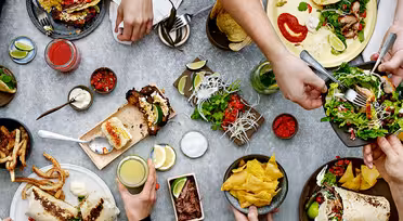 Ein Tisch voller verschiedener Tex-Mex-Gerichte, darunter Tacos, Salat und Nachos. Mehrere Hände greifen hinein und nehmen sich Essen. Beilagen wie Salsa, Limettenspalten und Getränke stehen verteilt auf dem Tisch.