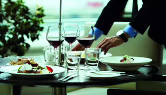 Eine Person im Anzug deckt einen formellen Esstisch mit Tellern voller Gourmet-Essen, Gläsern Rotwein und Wasser. Der Tisch ist elegant arrangiert, im Hintergrund steht eine kleine Pflanze, was an ein Restaurant erinnert.