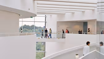 Ein moderner, geschwungener Innenraum mit weißen Wänden und einer Wendeltreppe. Die Menschen gehen auf einem oberen Gehweg mit Glasgeländer. Große Fenster bieten einen Blick auf die Bäume draußen und schaffen eine großzügige und luftige Atmosphäre.