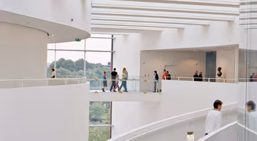 Ein moderner, geschwungener Innenraum mit weißen Wänden und einer Wendeltreppe. Die Menschen gehen auf einem oberen Gehweg mit Glasgeländer. Große Fenster bieten einen Blick auf die Bäume draußen und schaffen eine großzügige und luftige Atmosphäre.