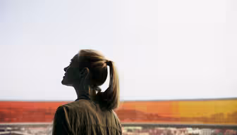 Eine Person mit Pferdeschwanz steht im Haus und blickt durch ein großes Fenster. Das Fenster hat farbenfrohe, orange-gelb getönte Bereiche und bietet einen Blick auf eine Stadtlandschaft unter bewölktem Himmel. Die Person ist im Profil zu sehen und trägt eine Jacke.