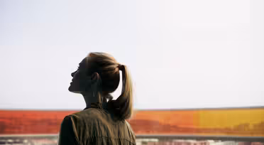 Eine Person mit Pferdeschwanz steht im Haus und blickt durch ein großes Fenster. Das Fenster hat farbenfrohe, orange-gelb getönte Bereiche und bietet einen Blick auf eine Stadtlandschaft unter bewölktem Himmel. Die Person ist im Profil zu sehen und trägt eine Jacke.