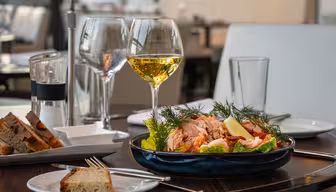 Ein gedeckter Tisch mit einem Salat mit Lachs, Zitronenspalten und Dill in einer blauen Schüssel. Daneben stehen Schwarzbrotscheiben, zwei Gläser Weißwein, eine Pfeffermühle und ein Glas Wasser. Im Hintergrund ist ein stilvoller Essbereich zu sehen.