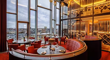 Luxuriöses Restaurant mit rot akzentuiertem Dekor und großen Fenstern mit Blick auf die Stadt. Runde Esstische sind mit Tellern und Gläsern gedeckt. Eine Wand mit Weinregalen und Hängelampen verleiht dem Ambiente Wärme und Eleganz.