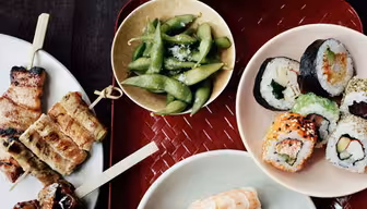 Verschiedene Sushi-Rollen, darunter Nigiri mit Garnelen und Thunfisch, werden auf Tellern angerichtet. Dazu gibt es gegrilltes Fleisch am Spieß und eine Schale Edamame. Das Ganze wird auf einem rot geflochtenen Tablett serviert, daneben stehen eine Schale Reis und Essstäbchen.